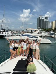 Group of smiling women raising their hands on the bow of a yacht at a sunny marina, surrounded by luxury boats and a modern high-rise skyline.