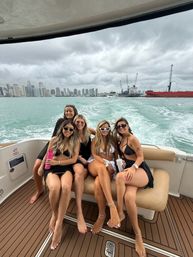 Five friends in swimsuits smiling on the back of a yacht, turquoise water trailing behind, Miami skyline and cargo cranes on the horizon under a dramatic cloudy sky