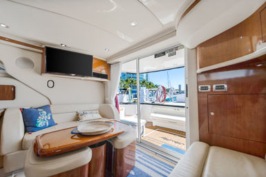 Bright luxury yacht salon with cream leather seating, glossy wood table and flat-screen TV, sliding glass doors opening to a sunny marina with palm trees and sailboats