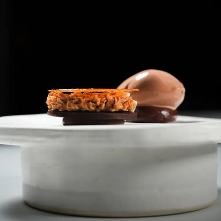 Chocolate plated dessert on a white marble pedestal — dark chocolate discs topped with a crunchy caramelized nut layer and thin brittle, paired with glossy chocolate ice cream and sauce against a black background.