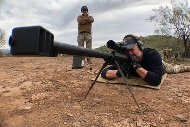 Shooter lying prone on an arid outdoor range aiming a long-range precision rifle on a bipod while an observer stands behind under a cloudy sky.