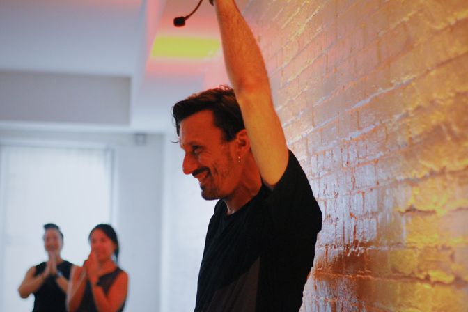 Smiling yoga instructor raises an arm against an exposed brick wall in a bright urban studio while students applaud