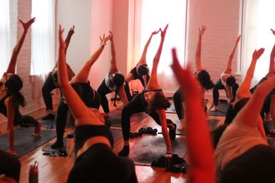 Energized group yoga class in a bright urban studio practicing triangle/side-angle stretches on mats with warm red lighting, wooden floors and water bottles nearby
