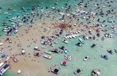 Aerial view of dozens of boats and pontoons anchored around a packed sandbar party, with crowds of people wading in shallow turquoise coastal water.