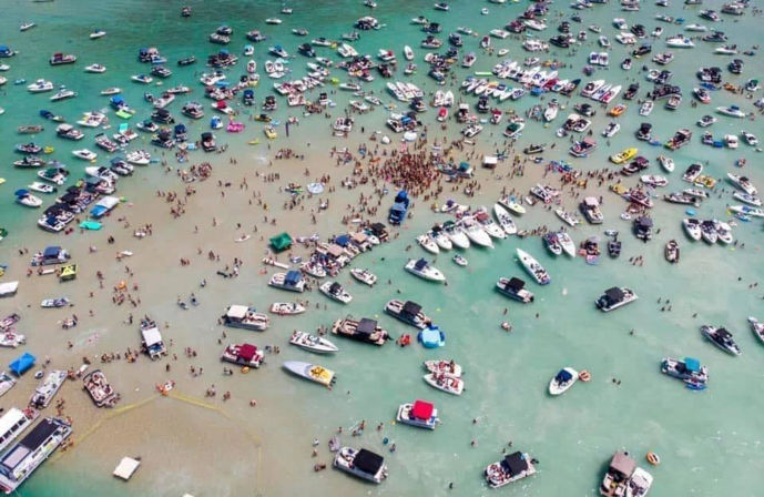 Aerial view of dozens of boats and pontoons anchored around a packed sandbar party, with crowds of people wading in shallow turquoise coastal water.