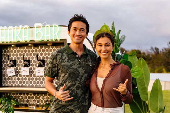 Two smiling people posing with shaka hand gestures in front of an outdoor tiki-style bar with a neon sign, hex-tile backsplash, drink taps and tropical plants at sunset.