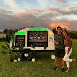 Two smiling people pose by a white mobile trailer bar with neon signs, tropical potted plants, beer taps and hex tile shelving on a grassy lawn at sunset