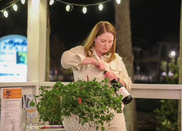 Person uncorking a bottle of wine on a string-lit outdoor patio at night, with a lush potted plant and a wine glass on the table nearby