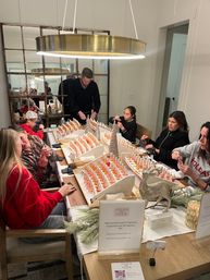 Family and friends gathered around a modern dining table making custom fragrances at a holiday workshop, rows of small glass scent bottles, festive greenery and a gold pendant light