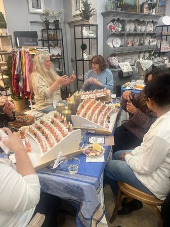 Women at a cozy boutique perfume sampling workshop, seated around a table with rows of small fragrance bottles, candles, snacks, and home-decor shelves in the background