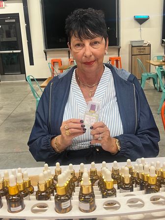 Woman at an indoor market table showcasing a small perfume bottle, rows of amber dropper bottles with gold caps on display, colorful metal cafe chairs and a 'thank you' bin in the background.