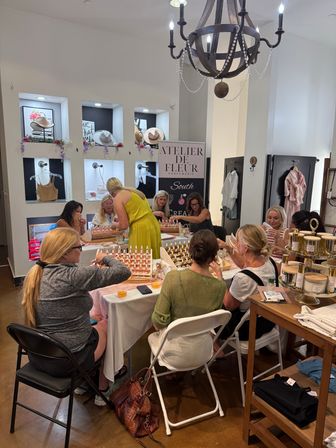 Small-group perfume-blending workshop in a bright boutique — women sampling fragrance vials at tables surrounded by candles, clothing displays and a decorative chandelier.