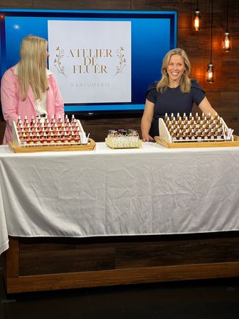 Two smiling hosts at a TV studio table showcasing a boutique perfumery product display — rows of small perfume bottles with droppers on wooden trays, white tablecloth, logo on screen and warm pendant lights.