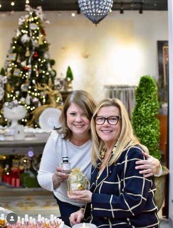 Two smiling women in a festive holiday boutique holding a decorative gold ornament and a small perfume bottle, with a lit Christmas tree and gift displays in the background