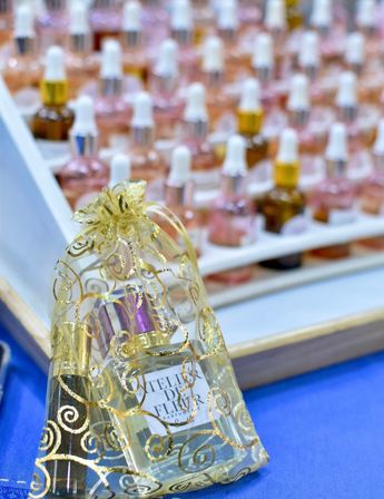 Gold organza gift pouch with shimmering swirls holding a small perfume bottle and cosmetics on a blue table, with rows of pink and amber dropper bottles blurred on a display tray in the background