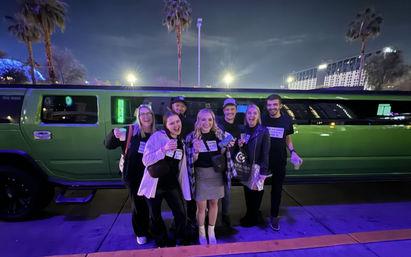 Group of seven friends smiling and holding drinks in front of a bright green stretch limo at night with palm trees and city lights — festive nightlife scene