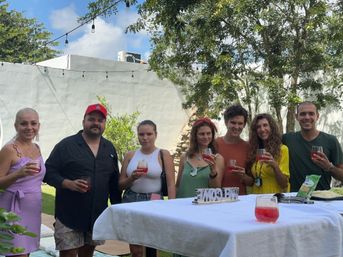 Group of seven people holding red cocktails at a sunny backyard garden party with string lights, trees, a white patio wall, and a 'WELCOME' sign on a covered table.