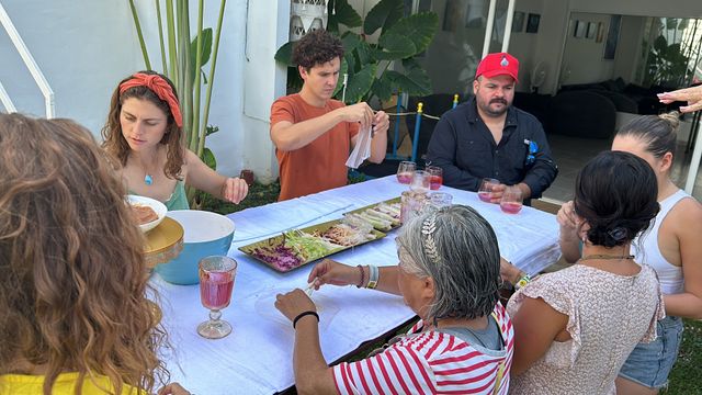 Group of people at an outdoor patio table sharing a colorful family-style meal — rectangular platter of fresh salad and wraps, pink drinks in goblets, bowls and napkins, tropical plants in the background.