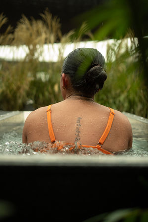Rear view of a person in an orange swimsuit sitting in an outdoor hot tub, water bubbling around their shoulders, droplets on skin, hair in a neat bun and a vertical script tattoo along the upper spine, framed by tall grasses and greenery.