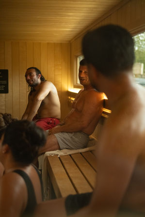 Adults relaxing in a wood-paneled indoor sauna, sitting on benches with towels under warm amber lighting for a casual wellness spa vibe.
