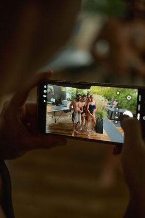 Hands holding a smartphone taking a photo of four friends in swimsuits posing on a wet wooden backyard deck by a hot tub and lush greenery