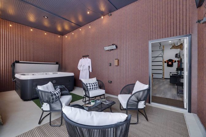 Cozy covered patio spa area with a large hot tub, four black rattan lounge chairs with white cushions around a low coffee table, string lights overhead, robes hanging on the wall, and a doorway into a garage-style room with a mounted deer head.
