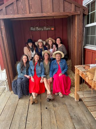 Eight women in sundresses, denim jackets and wide-brim hats smiling while seated on hay bales inside a rustic red barn doorway — casual country-style group portrait.