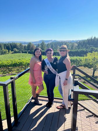 Three women on a sunny winery terrace overlooking grapevines and rolling hills, one wearing a 'Birthday Queen' sash and holding a wine glass during an outdoor celebration.
