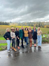 Eight women posing on wet pavement in front of a rolling autumn vineyard with colorful vines and a dramatic overcast sky