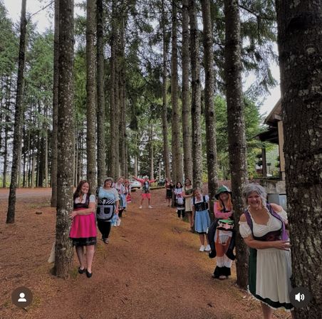 Playful group in dirndl-style dresses lined up peeking from behind tall pine trees along a woodland path, with a cabin and parked car visible in the forest background.