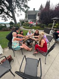 Group of women toasting with small glasses around a metal patio table on a landscaped lawn in front of a Victorian-style house