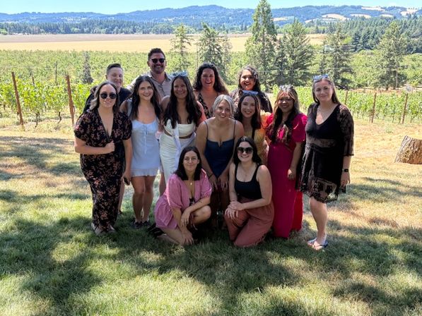 Cheerful group of friends posing for a sunlit summer photo in a vineyard, with rows of grapevines, trees and rolling hills under a clear blue sky.