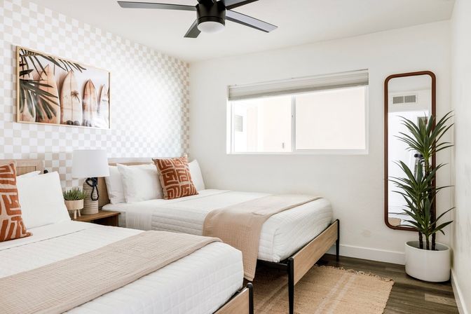 Sunlit coastal-inspired twin bedroom with two white quilted beds, beige throws and terracotta patterned pillows, checkered accent wall with surfboard artwork, black ceiling fan, tall potted plant by a full-length mirror, and a window letting in natural light.