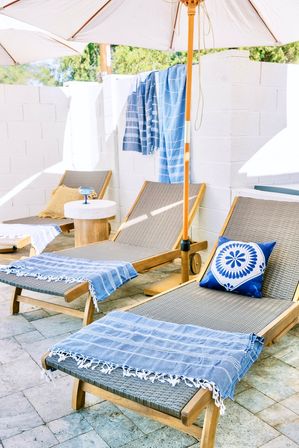 Sunlit outdoor patio with wooden sun loungers on stone tiles, blue-striped towels draped over the chairs, a blue patterned throw pillow on one lounger, and white umbrellas providing shade.