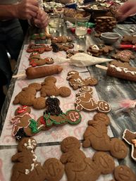 Festive holiday gingerbread cookie decorating scene on a kitchen counter with iced gingerbread men, piping bags, sprinkles, drinks, and hands adding icing.