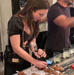 Person piping black icing onto gingerbread cookies at a cozy cookie-decorating gathering on a marble countertop with drinks and a smartphone nearby.