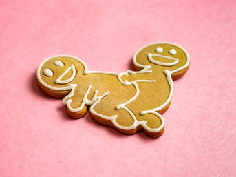 Playful iced gingerbread cookie shaped like two smiling figures joined together, white icing details on a pink background, close-up food photo