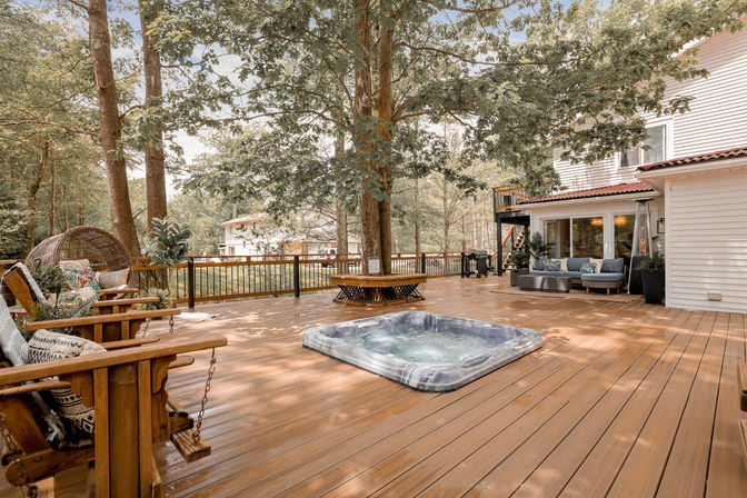 Spacious residential wooden deck with inset hot tub, large shade trees and circular bench, cozy outdoor sofa and swing chairs beside a white house — a relaxing backyard patio.