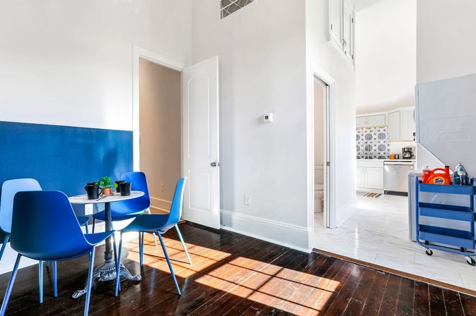 Bright sunlit modern apartment dining nook with four blue chairs around a small round table with potted succulents, dark hardwood floors, half-blue painted wall, and an open-plan white kitchen with patterned backsplash and laundry cart.