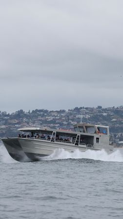 Silver passenger catamaran ferry cutting through choppy bay waters with passengers under a covered cabin and a foggy coastal hillside of homes in the background