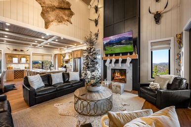 Cozy mountain-lodge open-plan living room in modern farmhouse style, decorated for Christmas with a frosted tree, stockings on a lit fireplace, leather sofas, round coffee table, mounted horns and a window framing a mountain view.