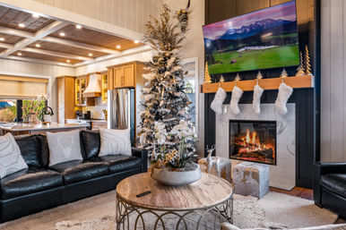 Cozy modern living room with a decorated Christmas tree, lit fireplace with four fluffy stockings on the mantel, leather sofa, round coffee table with festive centerpiece, and open kitchen with wood‑beam ceiling