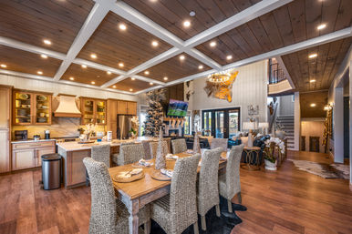 Open-concept rustic mountain-lodge interior with coffered wood ceiling, farmhouse dining table with woven rattan chairs, kitchen island and stainless appliances, decorated Christmas tree and cozy living area.