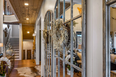 Modern farmhouse foyer with distressed multi-pane mirror doors adorned with a metallic wreath, wood-plank ceiling, hardwood floors with cowhide rug, decorative console and staircase opening to a cozy living area.