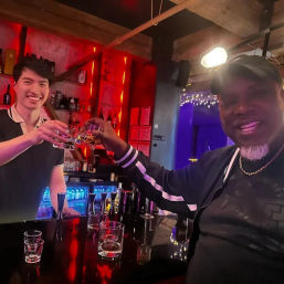 Two people toasting at a neon-lit cocktail bar — smiling bartender and patron clinking whiskey glasses over a glossy black counter with backlit bottles and festive lights.