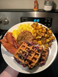 Hearty breakfast on a white plate: blueberry waffles with burst berries, crispy bacon, fluffy scrambled eggs, golden home fries and a sausage patty in front of a stovetop.