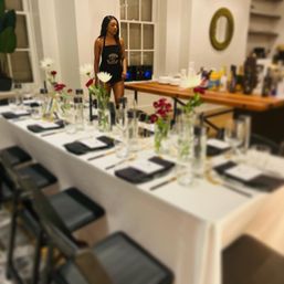 Modern dining room set for an intimate dinner: long white-tablecloth table with black napkins, glassware and red-and-white floral vases, while a woman in a black apron stands by a wooden serving table with bottles.