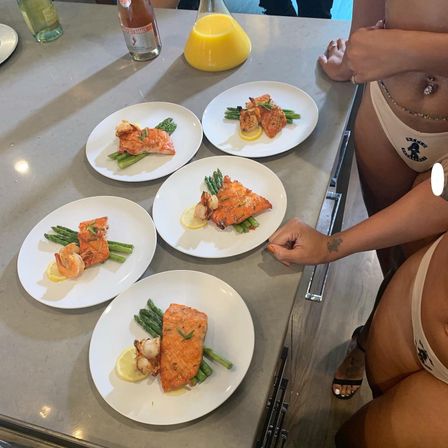 Five white plates on a kitchen island each with grilled salmon fillet on asparagus, a lemon slice and seared shrimp, with a carafe of orange juice and people in swimwear at the counter — summer seafood meal.