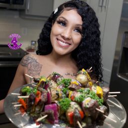 Smiling woman with curly hair in a kitchen holding a platter of colorful beef and vegetable skewers with bell peppers, red onion, potatoes and parsley.