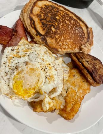 Classic American breakfast plate: golden pancakes, two fried eggs with black pepper, crispy hash browns, bacon strips and a sausage patty.